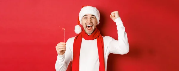 Concept of christmas, winter holidays and celebration. Portrait of excited handsome man, raising hand up and holding sparkler, wishing happy new year, standing over red background — Stock Photo, Image