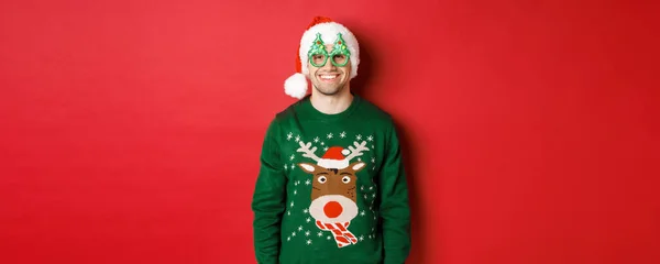 Retrato de hombre guapo en jersey de navidad verde, sombrero de santa y gafas de fiesta, sonriente feliz, de pie sobre fondo rojo —  Fotos de Stock