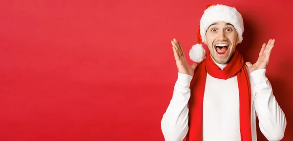 Концепция Рождества, зимних праздников и празднования. Изображение красивого мужчины, удивлённого новогодним промо-предложением, улыбающегося удивлённого, стоящего на красном фоне — стоковое фото