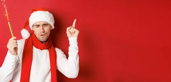 Begreppet jul, vinter semester och fest. Attraktiv kille njuter av nyårsfest, dans med glitter, bär tomte hatt och halsduk, står över röd bakgrund — Stockfoto