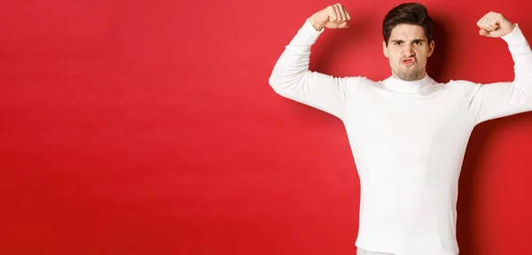 Portret van knappe en grappige man in witte trui, flex biceps en kijken aangemoedigd, met sterke spieren, staande over rode achtergrond — Stockfoto