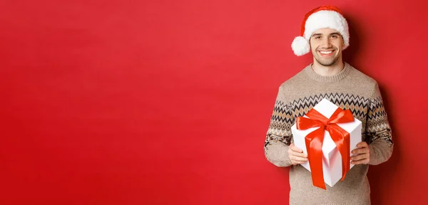 Imagem de homem barbudo bonito em camisola de inverno e chapéu de santa, segurando presente de Natal e sorrindo, tem um presente embrulhado com fita, de pé sobre fundo vermelho — Fotografia de Stock
