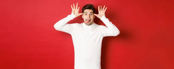 Porträt eines lustigen kaukasischen Typen, der Zunge zeigt und Gesichter macht, weißen Pullover trägt und vor rotem Hintergrund steht — Stockfoto