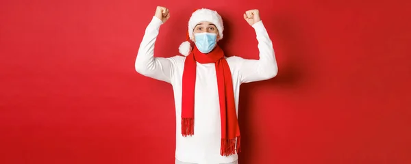 Covid-19, noel ve salgın sırasında tatil kavramı. Noel Baba şapkası ve tıbbi maske takmış mutlu bir adamın portresi, mutlu ve yeni yılı kutluyor, kırmızı arka planda duruyor. — Stok fotoğraf