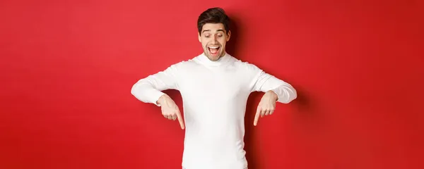 Porträt eines glücklichen, gut aussehenden Mannes, der mit dem Finger nach unten zeigt und die Werbung mit aufgeregtem Lächeln betrachtet, vor rotem Hintergrund stehend — Stockfoto