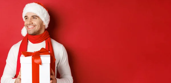 Концепция зимних праздников, Рождества и образа жизни. Крупный план привлекательного парня в шляпе Санты и шарфе, улыбающегося и выглядящего слева, держа в руках подарок на Новый год, стоящего на красном фоне — стоковое фото