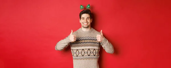 Begrip wintervakantie, kerstmis en viering. Vrolijke bebaarde man in trui, met duimen omhoog in goedkeuring en glimlachen, genieten van nieuwjaarsfeest, rode achtergrond — Stockfoto