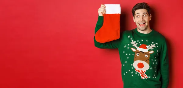 Yeşil süveterli, şaşırmış ve eğlenmiş bir adamın görüntüsü. Hediyelerle dolu Noel çoraplarına bakıyor ve gülümsüyor. Kırmızı arka planda duruyor. — Stok fotoğraf