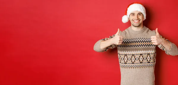 Imagem de homem atraente alegre em camisola de inverno e chapéu de Papai Noel, mostrando polegares para cima, celebrando o Natal e parecendo feliz, como algo, de pé sobre fundo vermelho — Fotografia de Stock