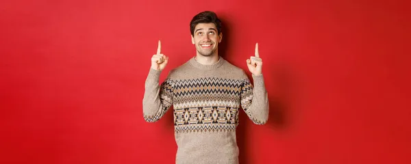 Muotokuva unenomainen komea kaveri ajatellut joulun vapaapäiviä, yllään talvi villapaita, osoittaa ja katselee ylös uusi vuosi promo tarjous, seisoo yli punainen tausta — kuvapankkivalokuva