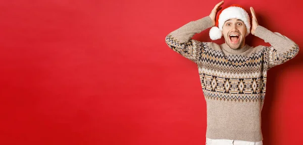 Imagem de jovem surpreso e feliz em chapéu de santa e camisola de Natal, recebendo presente incrível, de pé animado contra o fundo vermelho — Fotografia de Stock