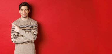 Noel süveteri giymiş yakışıklı bir adam resmi, yeni yılı kutluyor, mutlu bir şekilde gülümsüyor ve sol üst köşedeki fotokopi alanını işaret ediyor, kırmızı arka planda duruyor.