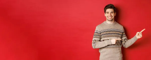 Obrázek atraktivního usmívajícího se muže, na sobě vánoční svetr, ukazující prsty vpravo a ukazující novoroční reklamu, stojící nad červeným pozadím — Stock fotografie