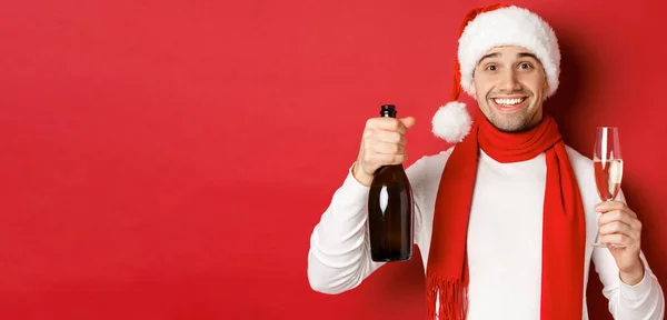 Pojęcie ferii zimowych, Bożego Narodzenia i stylu życia. Zbliżenie wesołego przystojniaka, trzymającego butelkę szampana i szklankę, wznoszącego toast za Nowy Rok i świętującego, czerwone tło — Zdjęcie stockowe