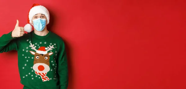 Concepto de año nuevo, covidio-19 y distanciamiento social. Joven sorprendido celebrando la Navidad, con máscara médica y sombrero de santa, mostrando el pulgar hacia arriba en la aprobación, fondo rojo — Foto de Stock