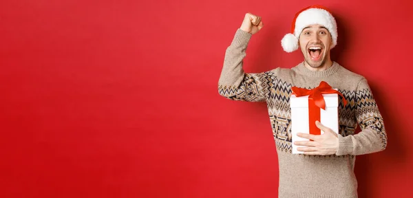 Εικόνα ενός χαρούμενου και ενθουσιασμένου όμορφου άντρα, που αναβιώνει το χριστουγεννιάτικο δώρο, σηκώνει τα χέρια ψηλά θριαμβευτικά και χαμογελά, γιορτάζει το νέο έτος, στέκεται πάνω από το κόκκινο φόντο στο καπέλο του Άη Βασίλη — Φωτογραφία Αρχείου