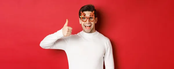 穿着派对眼镜和白色毛衣的英俊年轻人的特写镜头，展示大拇指向上看，享受新年假期，站在红色的背景上 — 图库照片