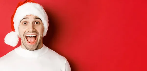 Fogalom a téli ünnepek, karácsony és ünneplés. Közelkép a meglepett jóképű férfi télapó kalap, hallani csodálatos újévi promóciós ajánlat, szemben a piros háttér — Stock Fotó