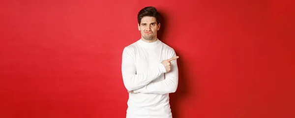 Doubtful and reluctant handsome man grimacing, pointing fingers right at something bad, standing in white sweater over red background — Stock Photo, Image
