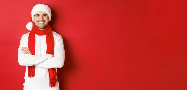 Bonito caucasiano com cerdas, vestindo chapéu de Papai Noel, cachecol e suéter branco, braços cruzados no peito e sorrindo feliz, de pé contra o fundo vermelho — Fotografia de Stock