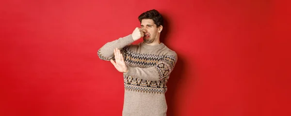 悪い匂いで何かを拒否し鼻を閉じて手を上げる冬のセーターのうんざりハンサムな男のイメージは、赤の背景の上に立って — ストック写真