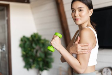 fit vücutlu aktif ve sağlıklı Asyalı kız evde spor yapıyor, dambılları kaldırıyor ve pazularına dokunuyor, oturma odasında egzersiz yapıyor.