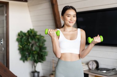Fitness Asyalı kız oturma odasında egzersiz yapıyor, dambılları kaldırıyor, spor etkinlikleri için etek giyiyor.