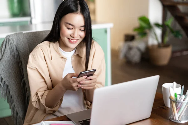 Asiática estudiante mirando el teléfono móvil, sentado en casa con el ordenador portátil, registrar curso en línea en el ordenador portátil, trabajando de forma remota — Foto de Stock