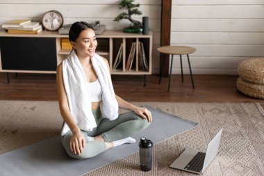 Spor ve yaşam tarzı konsepti. Gülümseyen genç Asyalı kadın evde yoga yapıyor, laptopta meditasyon kursunu takip ediyor, zen pozunda yer paspasında oturuyor.