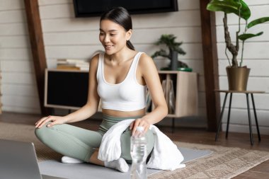 Meditasyon ve online sınıf konsepti. Evde uzaktan kumanda eğitimi alan genç bir kadın oturma odasında lastik paspasın üzerinde oturuyor ve yoga dersi için dizüstü bilgisayar kullanıyor.