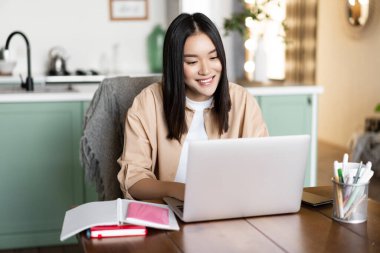 Görüntülü konuşma yapan bir kadın, bilgisayardaki online okul, dizüstü bilgisayardaki kameraya gülümseyen bir kadın. Evden uzakta okuyan bir kız, mutfakta kitap ve bilgisayarla oturuyor.