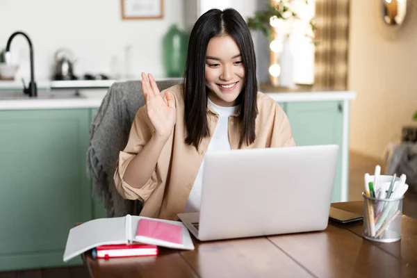 Imagen de chica asiática en videollamada, saludando con la mano en su video chat en el ordenador portátil y sonriendo. Estudiante o profesor saludando a la gente durante la clase remota en línea — Foto de Stock