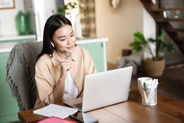 Genç Asyalı kadın ders çalışıyor, evden Webinar dinliyor, kulaklık takıyor ve bilgisayar ekranına bakıyor, internette ödev yapıyor.