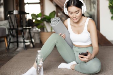 Su şişeli genç Asyalı kadın, evde spor yaparken müzik dinliyor ya da kulaklıkla podcast yapıyor, egzersiz yapmadan önce yerde oturuyor.