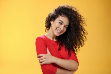 Kıvırcık saçlı, kıvırcık saçlı, başı öne eğik romantik bakışlı, romantik bakışlı, sarı arka planda kendine sarılan kadının belden yukarı pozu.