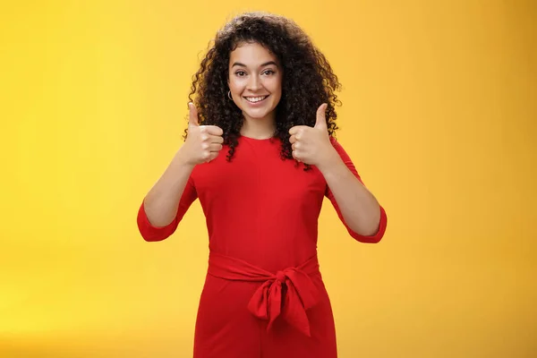 Excellent idea, nice job. Portrait of supportive delighted and happy charming female friend showing thumbs up as standing in red dress over yellow wall smiling giving positive reply, liking concept