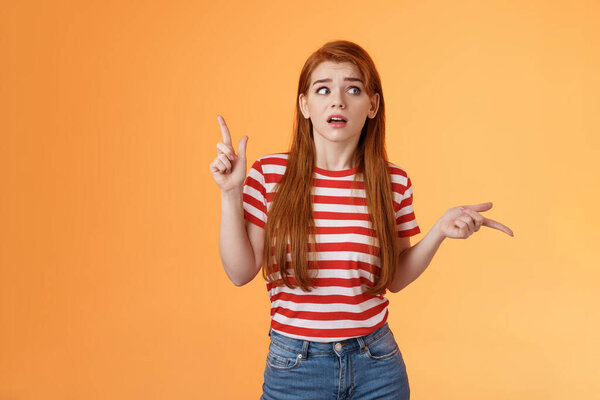 Complicated questioned redhead girlfriend trying understand what happening, which way go, look around, pointing sideways up and right, frowning perplexed, taking hard decision, orange background