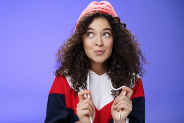 Tatlı ve aptal 20 'li yaşlarda, kıvırcık saçlı, sıcak kırmızı şapkalı, kıvırcık dudaklı, cilveli, sol üst köşesine şehvetle bakan ve mavi arka planda poz veren bir kız.