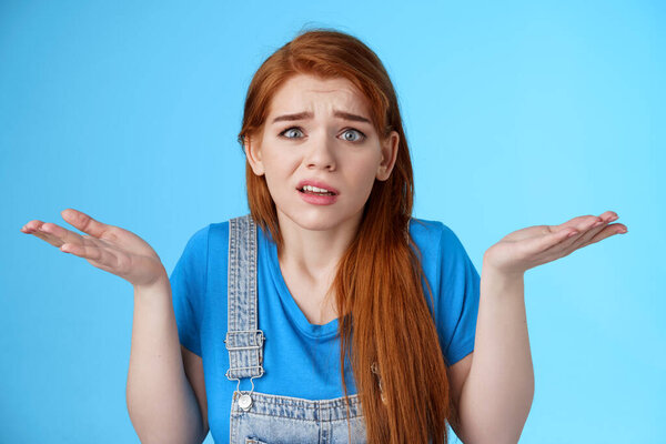 Perplexed uneasy stupified worried redhead woman shrugging, have no idea, frowning nervously, cannot understand what happened, stand clueless unsure, pose blue background