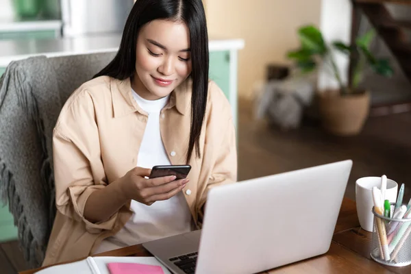 Imagen de una mujer asiática revisando mensajes telefónicos, trabajando en una computadora portátil desde casa, esperando una conferencia de trabajo, video chat — Foto de Stock