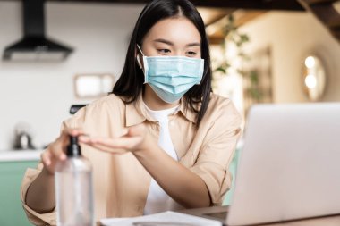 Tıbbi maskeli genç Asyalı kadın çalışma alanında oturuyor, online derse katılıyor ya da dizüstü bilgisayarda giyip el yıkamak için el dezenfektanı kullanıyor.