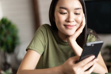 Yeşil tişörtlü gülümseyen Koreli kız cep telefonuna bakıyor, uygulamalı smth sipariş ediyor, akıllı telefon kullanarak evden alışveriş yapıyor.