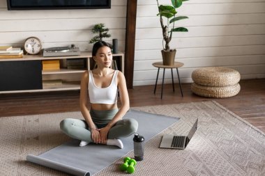 Asyalı fitness kızı evde dizüstü bilgisayarla antrenman yapıyor, spor uygulamasını bilgisayarda yapıyor oturma odasında paspasın üstüne oturuyor, etek giyiyor.