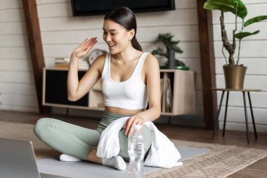 Asyalı fitness kızı lastik paspasa oturmuş yoga kursunda müşterileri selamlıyor ve dizüstü bilgisayarına el sallıyor.