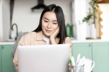 Gülümseyen Asyalı kadın rahatlıyor. Webinar izliyor, dizüstü bilgisayara bakıyor. Evde oturuyor.