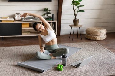 Genç Asyalı fitness kızı esneme hareketleri yapıyor, evde spor yapıyor, kollarını açıp dizüstü bilgisayara bakıyor, bilgisayardaki egzersiz videosunu takip ediyor.