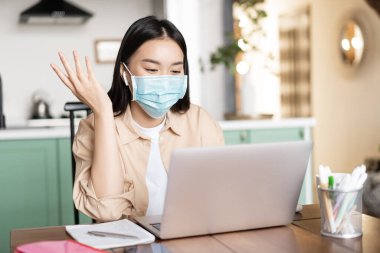 Evinden çalışan neşeli Asyalı kadın laptopla iletişim kuruyor, maske takıyor. Karantinadaki kız çevrimiçi derslerle çalışıyor