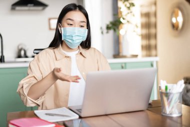 Genç Asyalı kız laptopta tıbbi maske takıp konuşuyor, evde oturuyor, çalışıyor ya da uzaktan çalışıyor.
