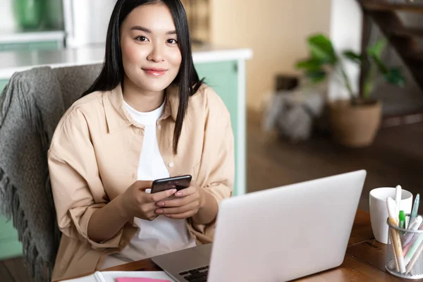 Sonriendo mujer asiática que trabaja con el ordenador portátil desde casa. Chica estudiante sosteniendo teléfono inteligente, sentado cerca de la computadora y mirando a la cámara — Foto de Stock