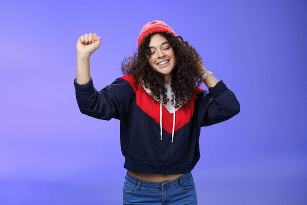 Girl delighted and carefree dancing over blue background with closed eyes and tender smile moving raised hands in rhythm of music, dreaming having positive mood wearing warm winter hat and sweatshirt
